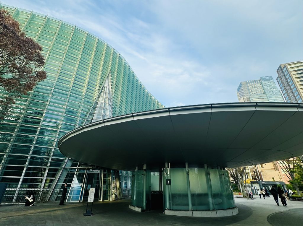 国立新美術館の正面玄関の写真。入口上部には巨大な円盤の雨避けがある。美術館本体はガラス張りで波打つような壁面の形をしている。