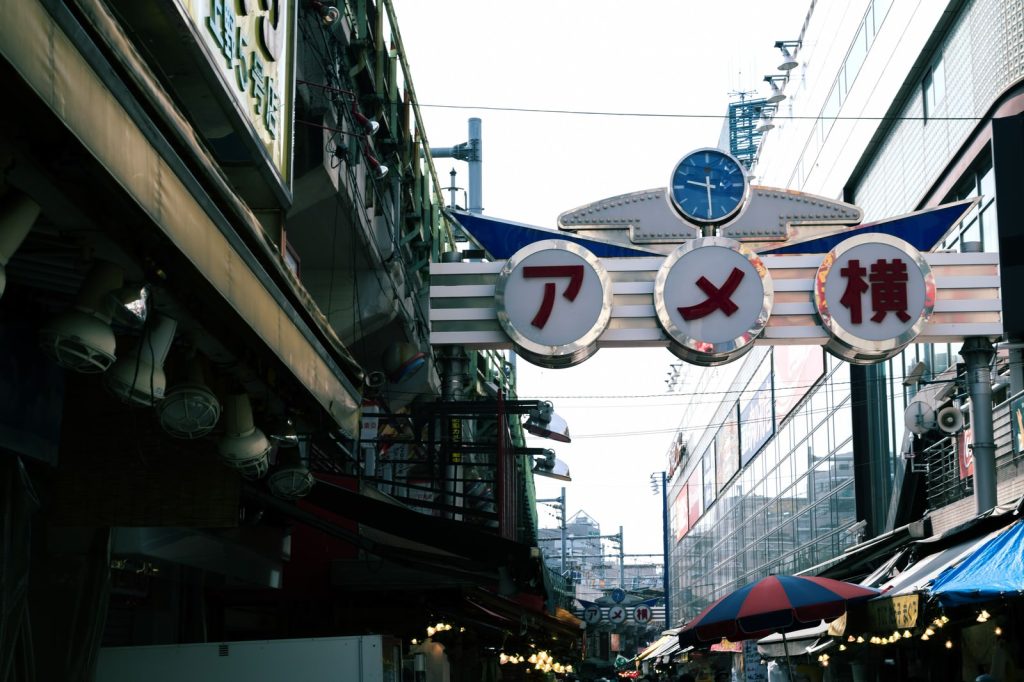 アメ横の商店街入口看板。左右の建造物に突っ張る形で設置されている。アメ横それぞれの字を丸く囲っていてその上に時計のあるデザイン。
