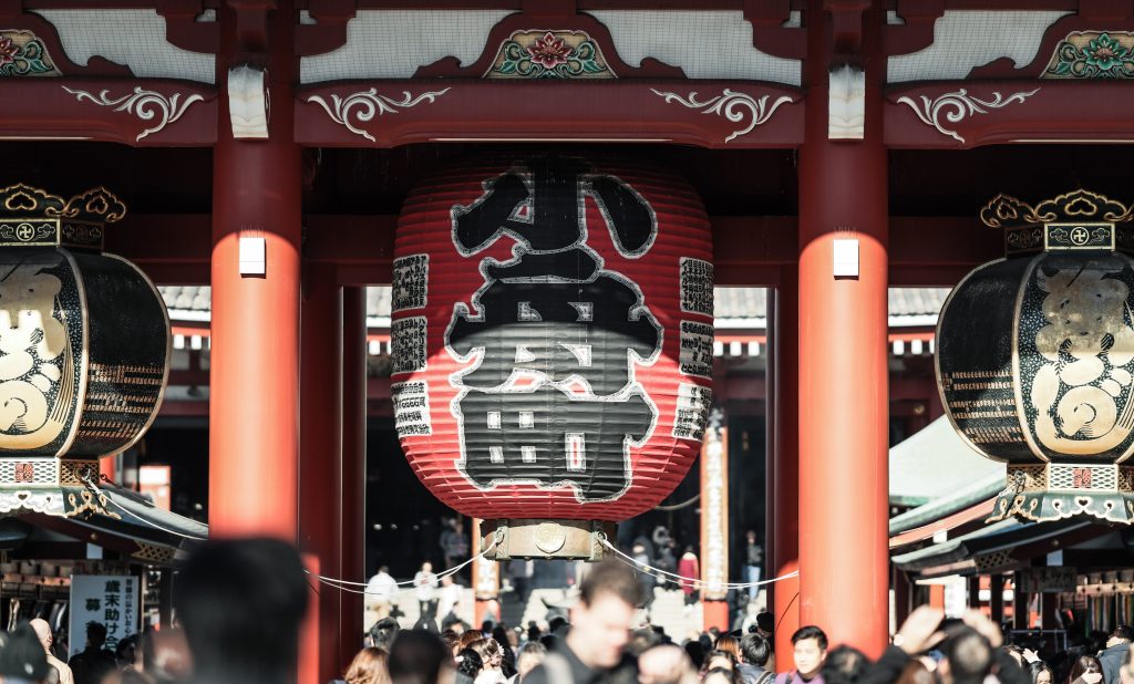 浅草寺の門の写真。奥には寺院があり、門を３つの柱が支えている。門の下には多くの人が通っており、頭上には大きな提灯が３つある。