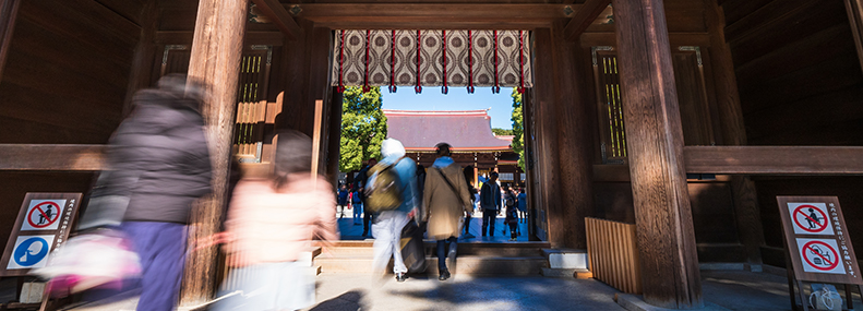 明治神宮門の写真。門の下を多くの人が通っている。奥には神宮がそびえ立つ。門の上側には帷がある。